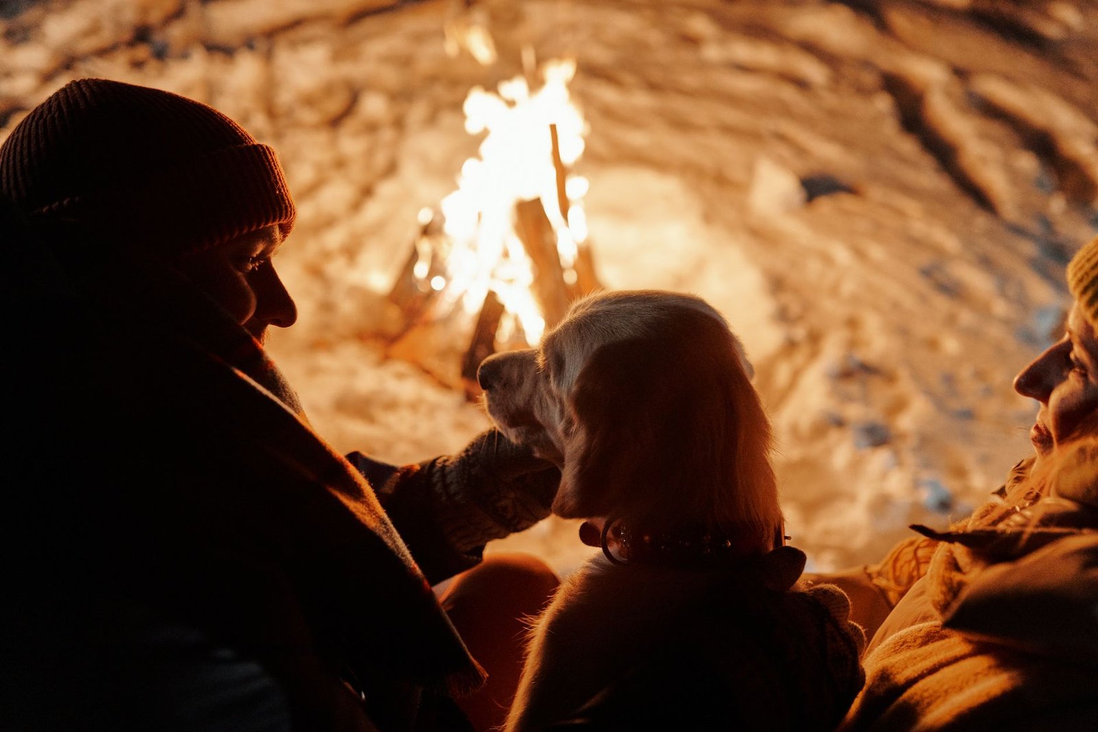 A couple enjoys a warm winter night by a bonfire, accompanied by their dog amidst a snowy setting.